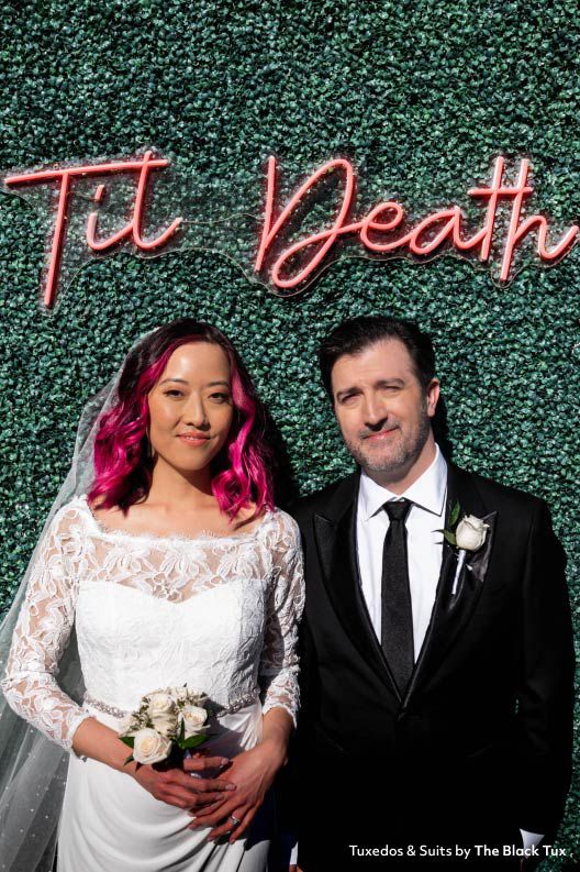 bride and groom standing in front of a neon sign that says til death