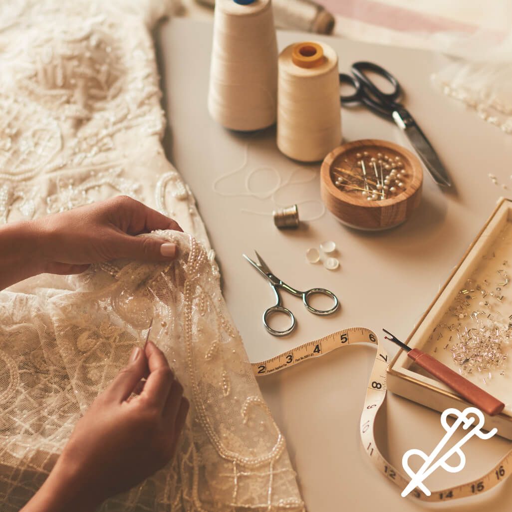 hands sewing a wedding dress on a table next to thread, containers of pins, scissors, and measuring tape