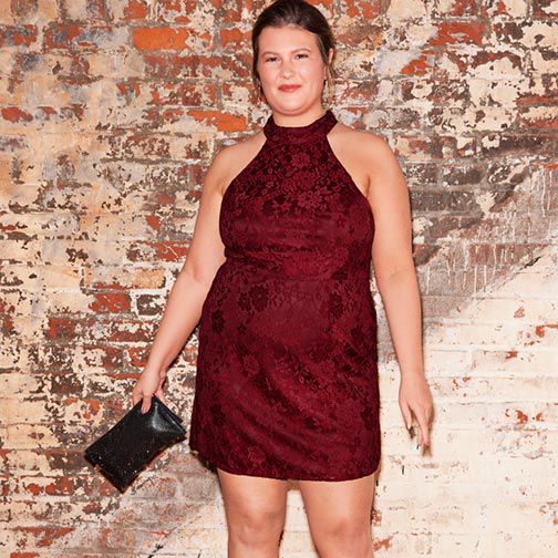 woman wearing a red occasion dress holding a handbag and standing against a brick wall