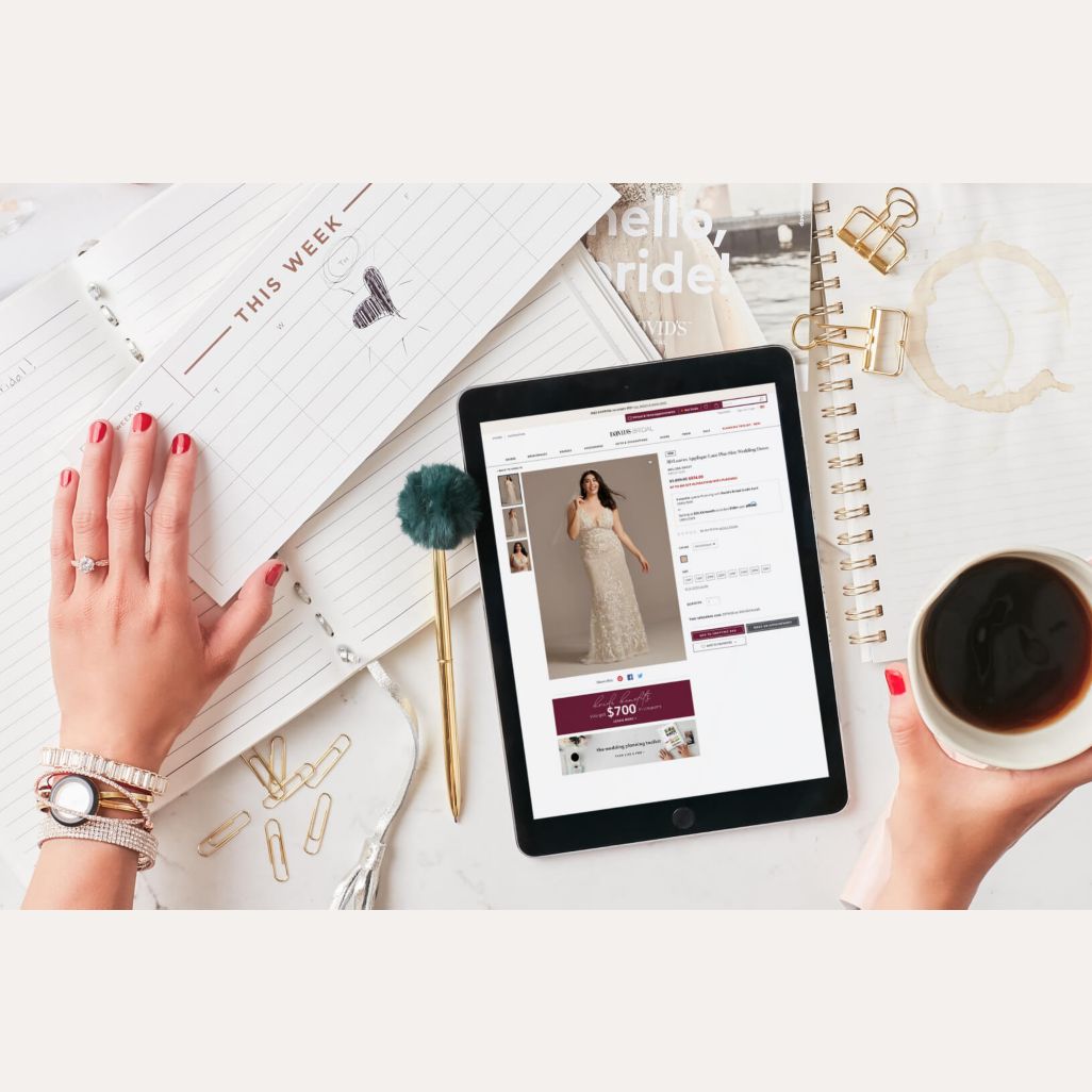 hand holding a cup of coffee looking at an image of a bride on an ipad on a table next to a calendar