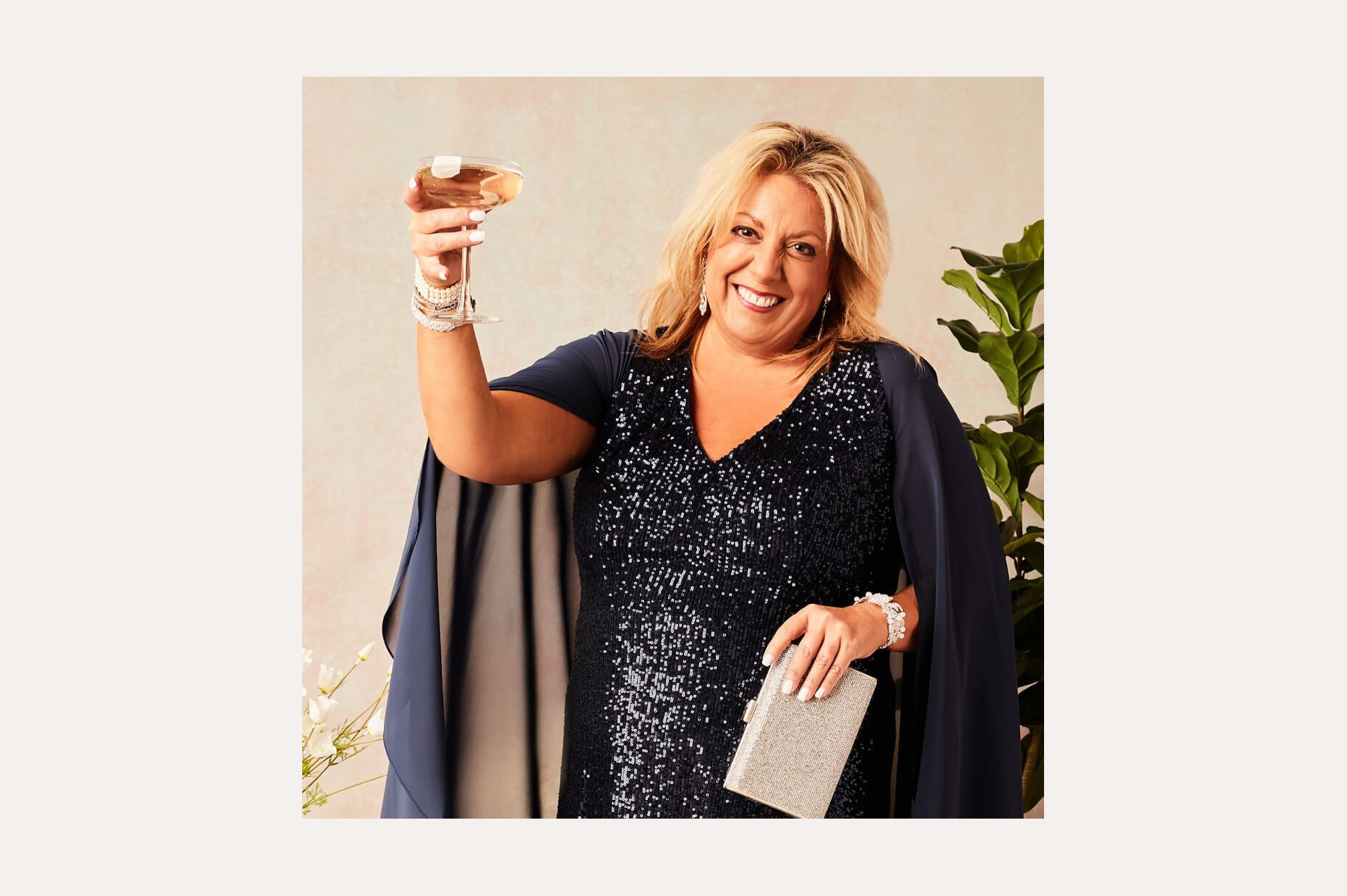 woman in a navy sequined dress and capelet smiling and holding up a glass of wine