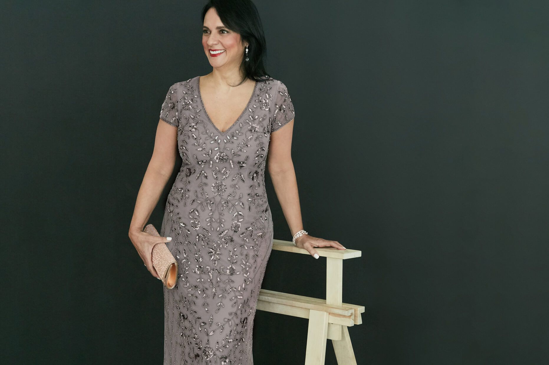 woman in silver beaded dress holding a clutch standing and leaning on a wood stand