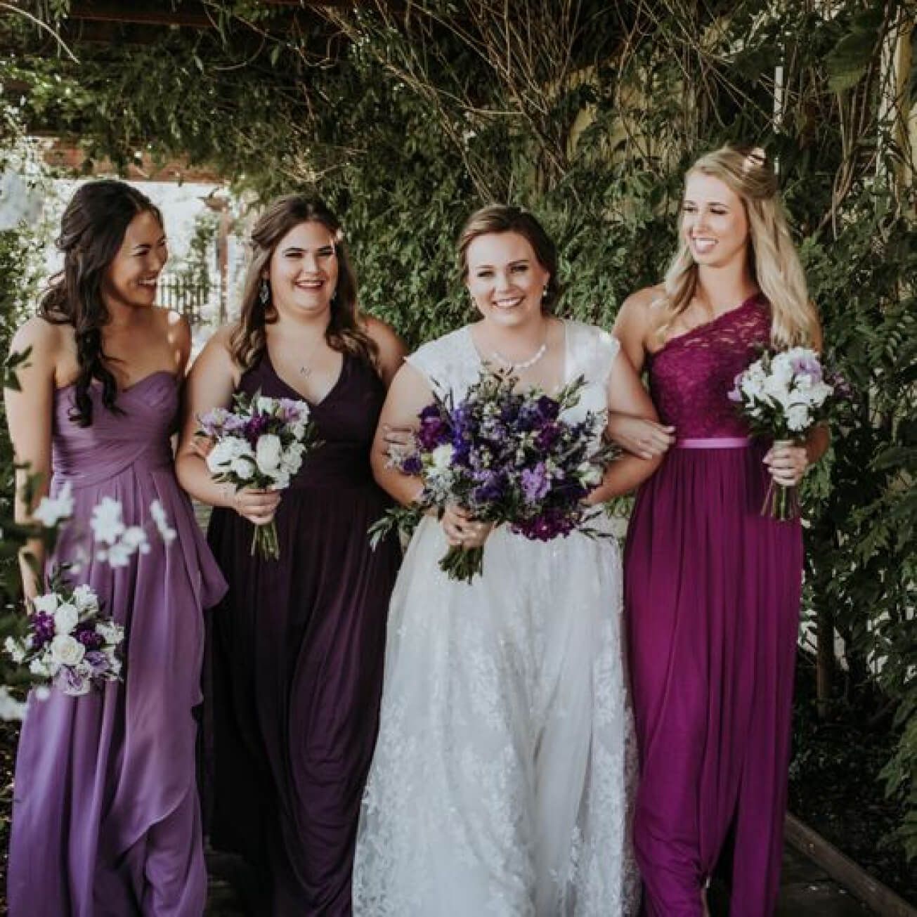 bride in a wedding dress walking with bridesmaids in different purple dresses holding flower bouquets